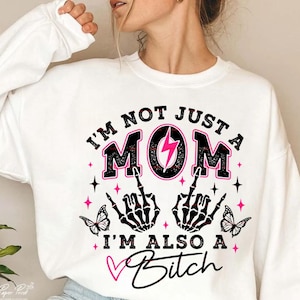 以下が含まれることがあります： 「I'M NOT JUST A MOM I'M ALSO A Bitch」の文字が入った白いスウェットシャツ。 ピンクの稲妻、中指を立てた黒い骸骨の手、蝶のデザインが含まれています。
