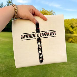 May include: A cream-colored fabric pouch featuring a black graphic of a cross with a distressed texture. The text "FATHERHOOD IS KINGDOM WORK" is printed horizontally across the cross, and "PROVERBS 22:6" is printed vertically along the base.