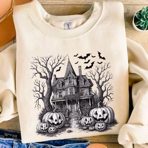 Pode incluir: Camisola creme com um design de Halloween a preto e branco. O gráfico inclui uma casa assombrada assustadora, abóboras de Halloween, árvores despidas e morcegos. Um artigo de vestuário festivo e sazonal.