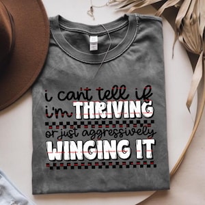 May include: Gray t-shirt with the text "i can't tell if i'm THRIVING or just aggressively WINGING IT" in black, red, and white. The shirt has a crew neck and short sleeves. A brown hat is partially visible in the background.