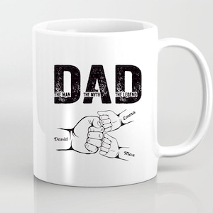 以下が含まれることがあります： 白いセラミックマグカップ。大きな黒い文字で「DAD」と書かれています。その下に「THE MAN, THE MYTH, THE LEGEND」という言葉があります。その下に、「David」、「Emma」、「Max」という名前の付いた手が描かれています。