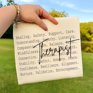 Op de afbeelding: Een rechthoekig, gebroken wit canvas met het woord "Therapist" in het zwart geschreven. Het canvas bevat woorden als "Healing", "Safety" en "Support" in een lettertype in typemachinestijl. Een hand met een gouden armband houdt het canvas vast.