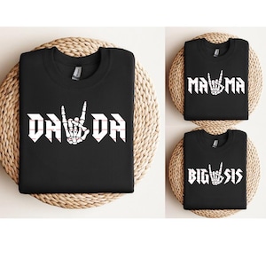 Op de afbeelding: Zwarte sweatshirts met witte tekst en een rock-and-roll handgebaar. Eén shirt zegt "DA DA", een ander zegt "MAMA" en de derde zegt "BIG SIS". De shirts worden gepresenteerd op geweven matten.