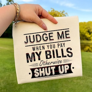 May include: A cream-colored canvas pouch with the text "JUDGE ME WHEN YOU PAY MY BILLS Otherwise SHUT UP" in black and red lettering. The pouch is held up against a background of green grass and a blue sky.