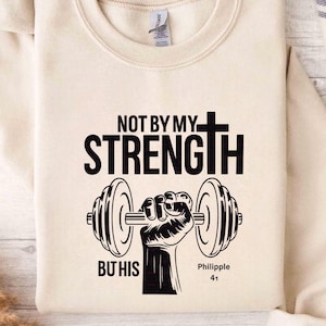 Könnte beinhalten: Cremefarbenes Sweatshirt mit dem Text "NOT BY MY STRENGTH" über einer Grafik einer Hand, die eine Langhantel hält. Darunter steht "BUT HIS" und "Philippians 4:13".
