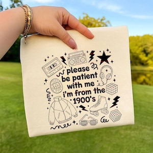 May include: Cream-colored canvas pouch with a black graphic design featuring 1900s-themed illustrations like a boombox, cassette tape, roller skate, and the text "please be patient with me I'm from the 1900's."