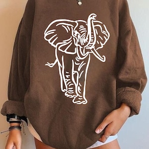 Puede incluir: Una sudadera marrón con un gráfico de elefante blanco. El elefante está dibujado en un estilo de contorno simple. La sudadera tiene cuello redondo y mangas largas con puños doblados. El estilo es informal.