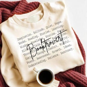 Op de afbeelding: Crèmekleurige sweatshirt met het woord "Booktrovert" in cursief, omringd door woorden als "Imagination" en "Reading". Een kopje koffie staat in de hoek, op een bordeauxrode deken.