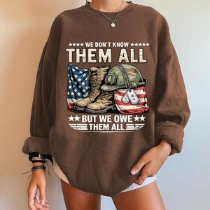 Könnte beinhalten: Braunes Sweatshirt mit einem Aufdruck von Militärstiefeln, einem Helm, Erkennungsmarken und der amerikanischen Flagge. Der Text lautet: "WE DON'T KNOW THEM ALL BUT WE OWE THEM ALL."