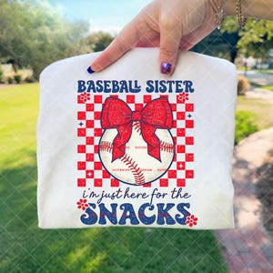 May include: White t-shirt with a baseball-themed graphic. The design features the text "BASEBALL SISTER" in blue, a red bow, and a baseball. Below, the text "i'm just here for the SNACKS" is in blue.