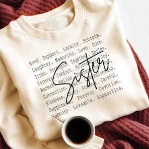 May include: Cream-colored sweatshirt with the word "Sister" in a handwritten font. The sweatshirt is printed with a list of words like "Bond, Support, Loyalty, Love, and Forever". A small cup of coffee sits on the folded garment.