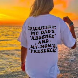 May include: A white t-shirt with the text "TRAUMATIZED BY MY DAD'S ABSENCE AND MY MOM'S PRESENCE" in black and red lettering. The shirt is worn by a person on a beach at sunset.
