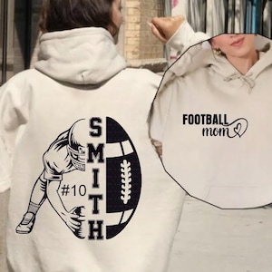 Op de afbeelding: Crèmekleurige hoodie met een zwarte afbeelding van een American footballspeler en een bal. Het woord "SMITH" staat verticaal op de bal gedrukt, samen met het nummer "10". Op de voorkant van de hoodie staat "FOOTBALL mom" met een hart.