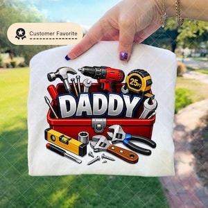 May include: White t-shirt featuring a red toolbox graphic filled with tools, including a hammer, drill, and measuring tape. The word "DADDY" is prominently displayed. A "Customer Favorite" badge is in the upper left corner.
