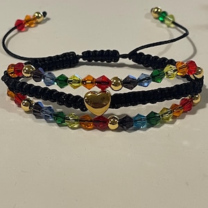 Pode incluir: Três pulseiras de macramé pretas com contas de cristal coloridas em arco-íris e detalhes dourados. A pulseira central apresenta um berloque de coração dourado. Nós deslizantes ajustáveis. Um acessório colorido e elegante.