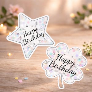 Pu&ograve; includere: Due adesivi decorativi con la scritta "Happy Birthday". Uno a forma di stella, l'altro a forma di quadrifoglio. Entrambi hanno un bordo bianco e sono riempiti di coriandoli pastello. Su una superficie di legno.