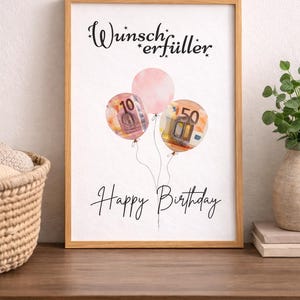 May include: A framed print with a light wood frame. The artwork features three balloons, two with currency designs and one pink. The text "Wunscherf&uuml;ller" and "Happy Birthday" are written in black script.