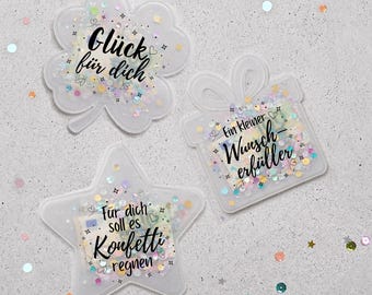 May include: Three clear, glitter-filled shapes: a four-leaf clover, a gift box, and a star. Each shape has black text. The clover says "Glück für dich", the gift box says "Ein kleiner Wunsch erfüller", and the star says "Für dich soll es Konfetti regnen".