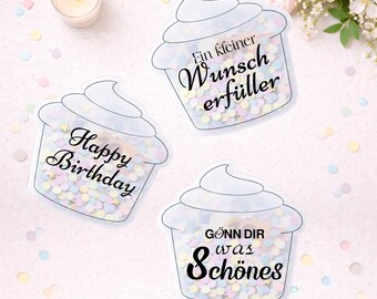 May include: Three cupcake-shaped cards with clear containers filled with confetti. One card says "Happy Birthday", another says "Ein kleiner Wunsch erfüller", and the third says "Gönn dir was Schönes". A lit candle and flowers are in the background.