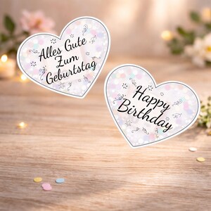 Pu&ograve; includere: Due adesivi a forma di cuore con sfondo bianco e design a coriandoli. Uno dice "Alles Gute Zum Geburtstag" e l'altro "Happy Birthday" in corsivo nero. Sono su una superficie di legno.