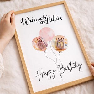 May include: A framed print with a light wood frame. The print features three watercolor balloons, two with currency designs and one pink. The text "Wunscherf&uuml;ller" and "Happy Birthday" are written in black script.