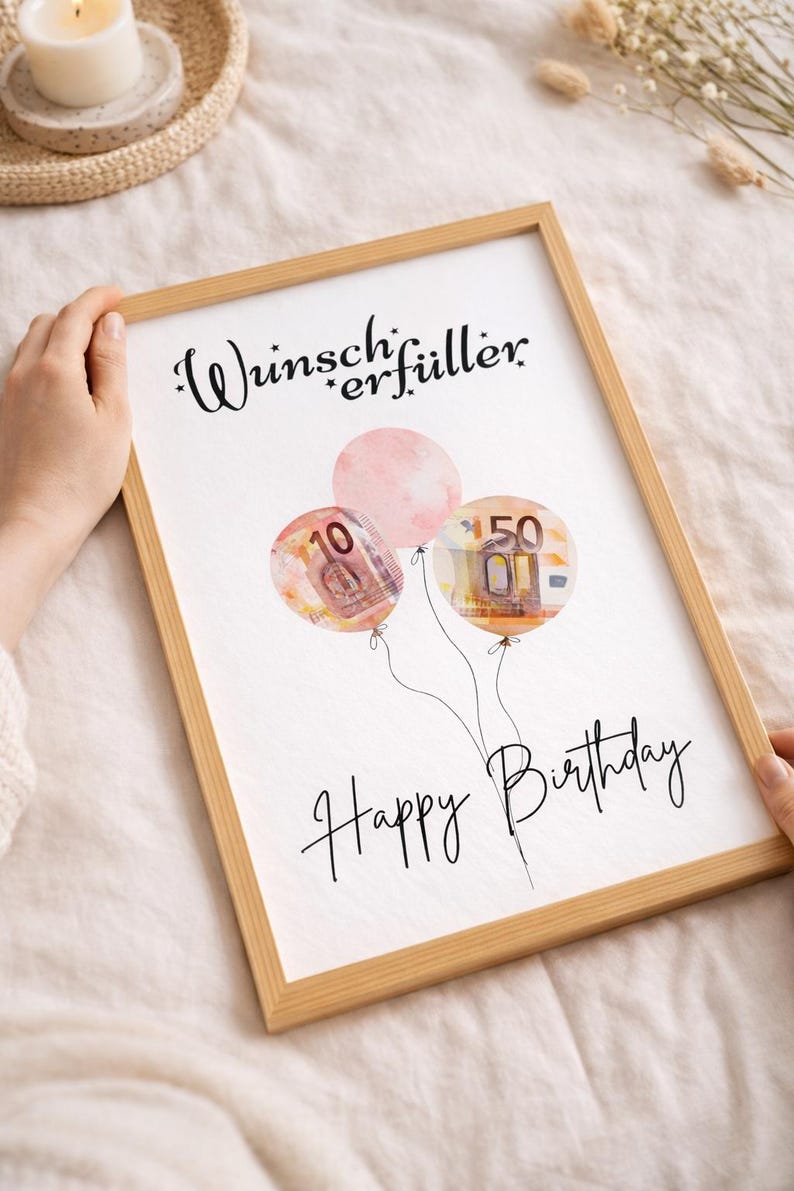 May include: A framed print with a light wood frame. The print features three watercolor balloons, two with currency designs and one pink. The text "Wunscherf&uuml;ller" and "Happy Birthday" are written in black script.