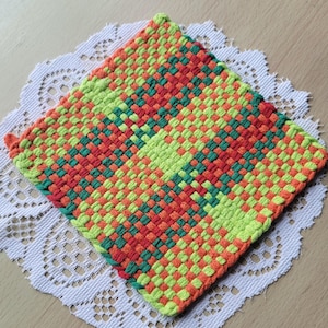 May include: A square, woven hot pad in a vibrant pattern of green, orange, and red. The potholder is made of a tightly woven material, with a loop for hanging. The hot pad is resting on a white, lacy doily.
