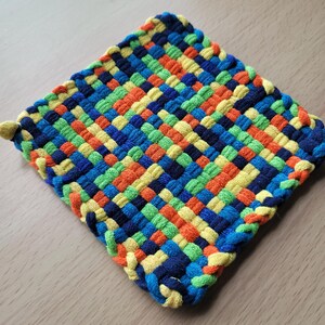 May include: A square, woven hot pad in a vibrant pattern of blue, green, orange, and yellow. The edges are neatly finished with a braided border. The handmade kitchen item is designed to protect surfaces from heat.