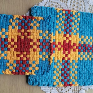 May include: Two square woven potholders featuring a geometric pattern in bright blue, golden yellow, and deep red. The textile construction displays a dense, interlocking weave with a consistent grid design across both pieces of kitchen linen.