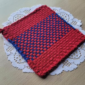 May include: A red and blue woven hot pad with a square design. The hot pad is made of thick, textured yarn and has a loop for hanging. It is placed on a white, lacy doily.