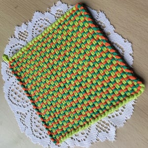 May include: A square, woven potholder in a vibrant pattern of green, orange, and yellow. The potholder is edged with green yarn and has a small loop for hanging. It rests on a white, lacy doily.