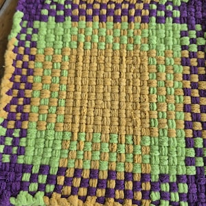 May include: A square potholder featuring a woven pattern in shades of purple, lime green, and mustard yellow. The design consists of a central mustard yellow square surrounded by alternating lime green and purple blocks in a grid-like textile weave.