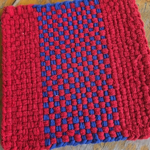 May include: A square potholder featuring a woven textile construction with a distinct pattern. The design consists of solid red borders on the left and right sides, with a central panel displaying a checkered pattern of alternating red and blue yarn.