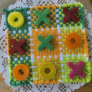 May include: A square woven potholder featuring a grid pattern in shades of green, yellow, and white. The surface is decorated with nine game pieces shaped like X's and O's in various colors including red, orange, yellow, and green.