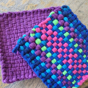 May include: Two square potholders made of woven fabric. One is a solid deep purple color with a small loop at the corner. The other features a multi-colored grid pattern in shades of bright blue, neon green, magenta, and dark purple.