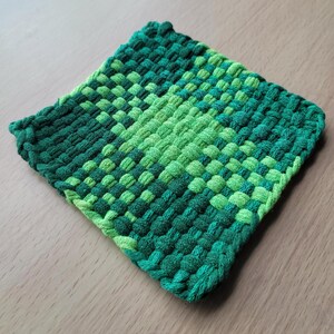 May include: A square, handwoven potholder in shades of green. The potholder features a woven pattern with varying shades of green, from light to dark. The edges are neatly finished with a thicker, darker green border. This kitchen accessory is designed to protect surfaces from heat.