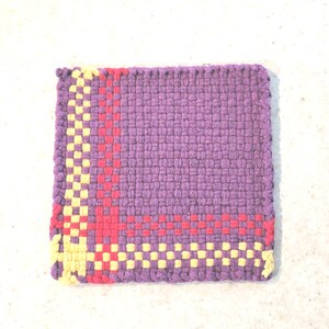 May include: A square, handwoven potholder in shades of purple, red, and yellow. The main body is a solid purple, with a border of alternating red and yellow squares. The potholder is made of woven fabric.