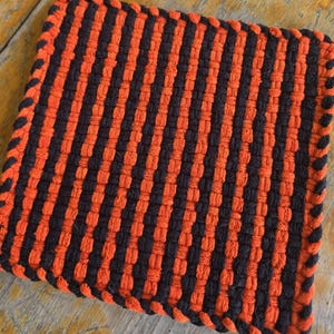 May include: A square potholder featuring a woven pattern of alternating vertical stripes in bright orange and black. The edges are finished with a braided border that alternates between the same orange and black colors, creating a textured surface.