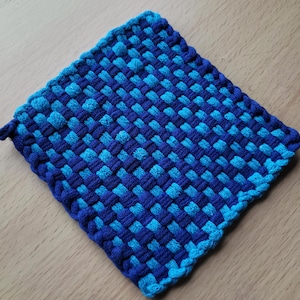 May include: A square, woven potholder in shades of blue. The design features a checkerboard pattern of dark and light blue threads. A small loop is attached to one corner for hanging.