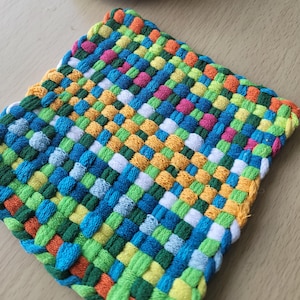 May include: A square, woven hot pad in a vibrant patchwork of colors. The design features a repeating pattern of interwoven threads in shades of blue, green, orange, yellow, pink, and white. The edges are finished with a yellow border.
