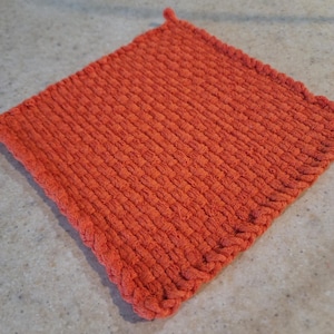 May include: An orange, square-shaped woven potholder. The potholder is made of a textured material and has a loop on one corner for hanging. The edges are finished with a decorative border.