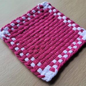 May include: A square, woven hot pad in a vibrant pink and white checkerboard pattern. The pink center is surrounded by a white border, and a small loop is attached for hanging. This handmade item is perfect for kitchen use.