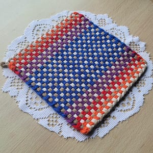May include: A square, woven hot pad with a gradient of colors, including red, orange, purple, blue, and tan. The hot pad is placed on a white, lacy doily. The woven design creates a textured surface.