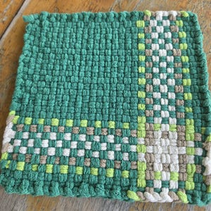 May include: A square woven potholder featuring a prominent dark green field with a vertical and horizontal band pattern. The accent stripes consist of alternating squares in white, light green, and tan, creating a grid-like texture throughout.