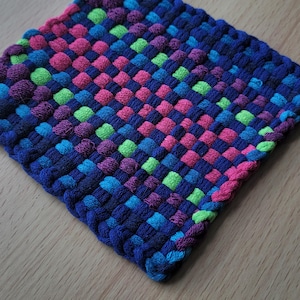 May include: A square, woven hot pad in a vibrant color scheme. The pad features a checkerboard pattern of blue, pink, purple, green, and blue squares. The edges are finished with a tightly woven border in a deep blue color.