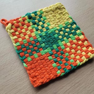 May include: A square, woven hot pad in a vibrant color scheme. The pad features a gradient design, transitioning from orange at the bottom to green and yellow at the top. A small loop is attached for hanging. The woven texture is clearly visible.