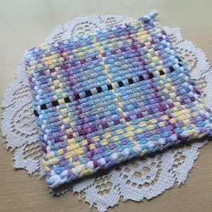 May include: A square, handwoven potholder in a plaid pattern. The potholder features a mix of colors including blue, purple, yellow, and gray. A black line runs across the center. The potholder is placed on a white lace doily.