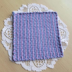 May include: A square, handwoven textile in shades of lavender and light blue, resting on a white lace doily. The woven piece has a textured appearance with vertical stripes of color. The doily has a delicate, floral pattern.