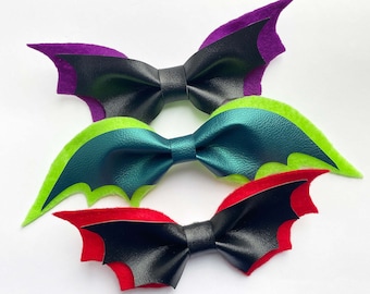 May include: Three bat-shaped hair bows in a row. The top bow is black with purple wings, the middle bow is teal with lime green wings, and the bottom bow is black with red wings. The bows are made of leather and felt.