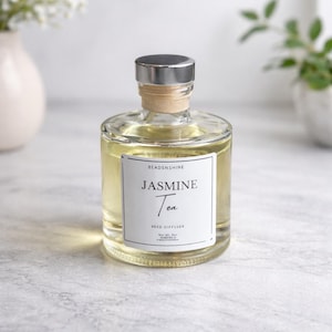 以下が含まれることがあります： 銀色のキャップとコルク栓が付いた、透明なガラス製のリードディフューザーボトル。ボトルには薄い黄色の液体が入っており、「Jasmine Tea」と書かれた白いラベルが付いています。ディフューザーは家庭用フレグランス製品です。